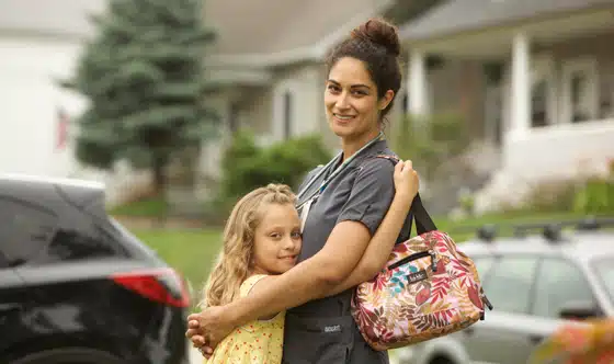 Young girl and woman in nurse attire with bag over shoulder are hugging in front of house with cars parked on the street