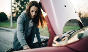 concerned woman in grey zip up hoodie peering under the hood of her red car
