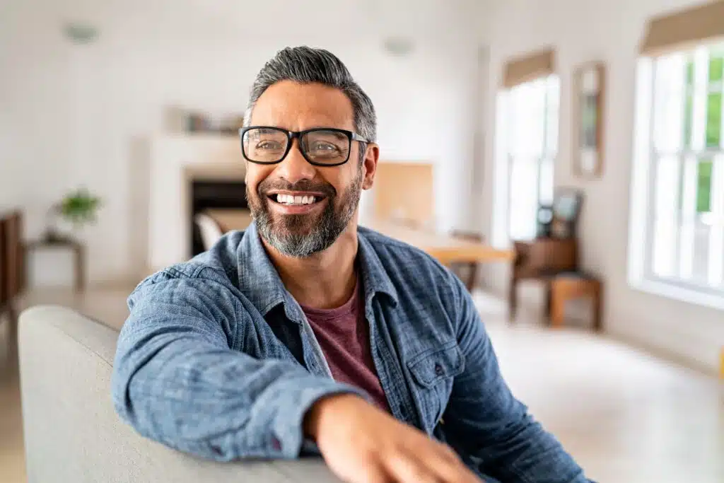 man in blue shirt sitting on sofa at home wearing glasses and smiling.
