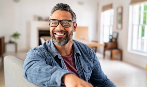 man in blue shirt sitting on sofa at home wearing glasses and smiling.
